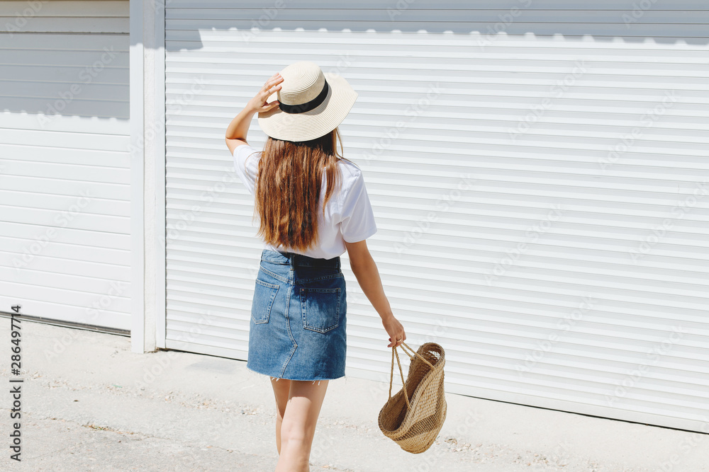 Young brunette woman wearing blue denim mini skirt, white t-shirt, wicker  straw bag and boater hat walking near white roller door. Trendy casual  summer or spring outfit. Street fashion. Back view. Stock