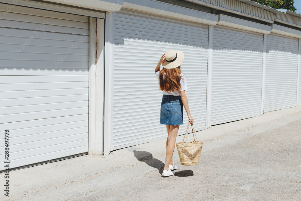 Young brunette woman wearing denim mini skirt, white t-shirt, sneakers, wicker straw bag, boater hat walking near white roller door. Trendy casual summer or spring outfit. Street fashion. Back view. Young brunette woman wearing denim mini skirt, white t-shirt, sneakers, wicker straw bag, boater hat walking near white roller door. Trendy casual summer or spring outfit. Street fashion. Back view.