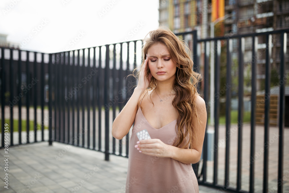 A beautiful girl is suffering from a headache and holding medicines in her hand while standing on the street A beautiful girl is suffering from a headache and holding medicines in her hand while standing on the street
