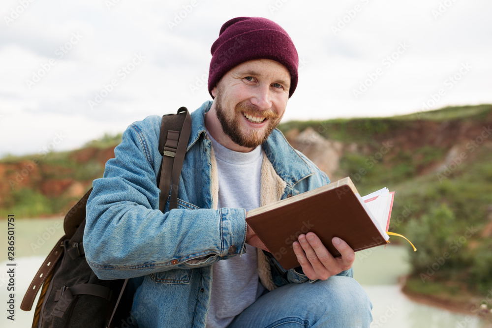Man traveler with a backpack on his shoulders sitting outdoors writing notes in a notebook about the observation of nature