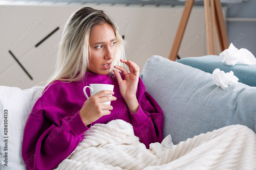 Concept of suffering from terrible pain. Beautiful, sad, upset, unhappy, troubled, weak woman with blonde hair, wearing casual clothes is sitting on a sofa and taking a pill Concept of suffering from terrible pain. Beautiful, sad, upset, unhappy, troubled, weak woman with blonde hair, wearing casual clothes is sitting on a sofa and taking a pill