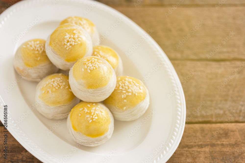 Chinese flaky pastry or moon cake filled with sweet mung bean paste and salted egg yolk on white plate decorated with white sesame in top view with copy space on wood table. Homemade bakery concept. Chinese flaky pastry or moon cake filled with sweet mung bean paste and salted egg yolk on white plate decorated with white sesame in top view with copy space on wood table. Homemade bakery concept.