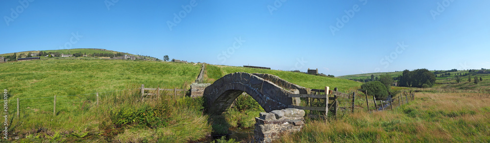 a panoramic view of an ancient stone packhorse bridge crossing a stream in west Yorkshire dales countryside with the village of colden in the background