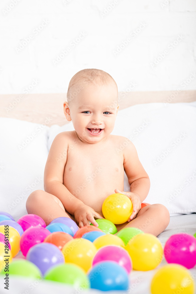 Cute little baby sitting on bed, playing with colored balls and laughing Cute little baby sitting on bed, playing with colored balls and laughing