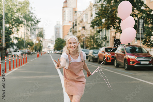 Happy cute blonde girl in bright clothes and balloons in hand stands on cityscape background, looks into camera and smiles. Blonde teenager with posing on the road with balloons, shows a tin of peace