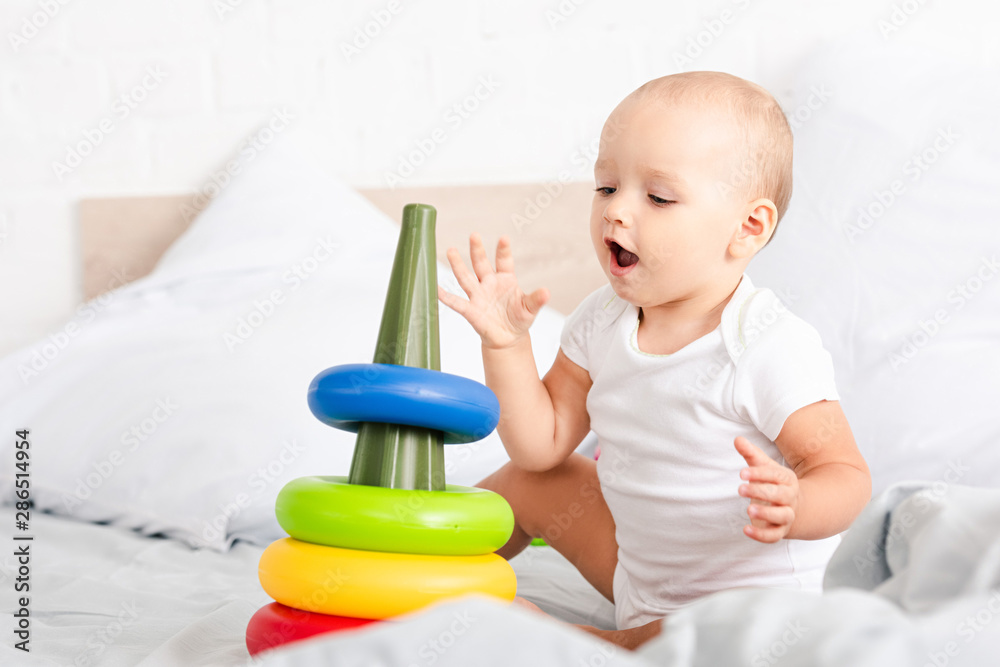 Cute little child in white clothes sitting on bed and playing with toy pyramid Cute little child in white clothes sitting on bed and playing with toy pyramid