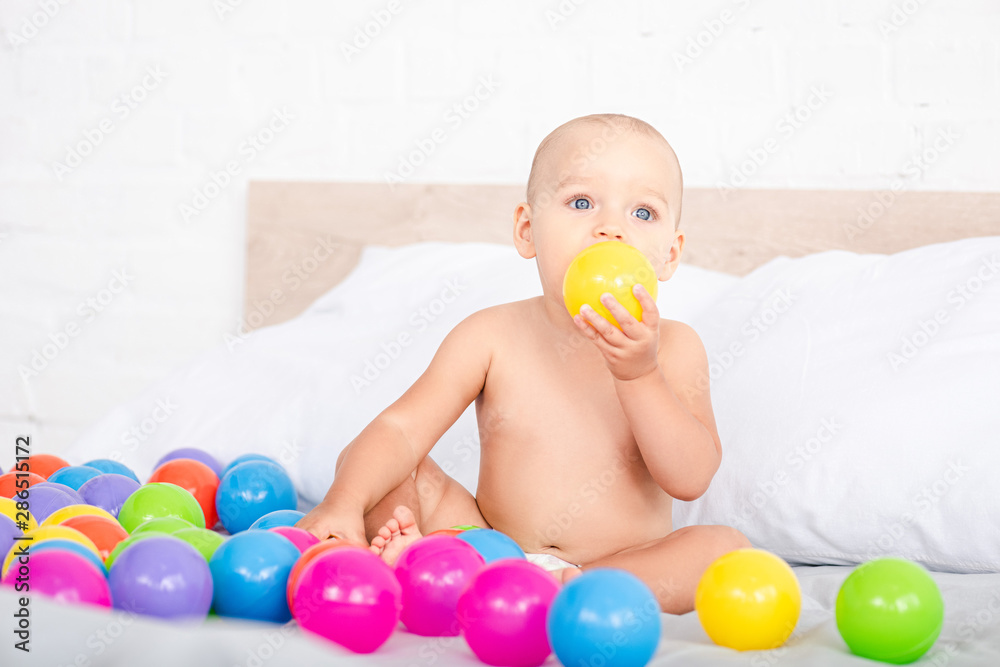Cute little baby sitting on bed and taking yellow ball into mouth Cute little baby sitting on bed and taking yellow ball into mouth
