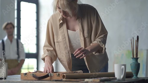 Female painter choosing painting tools at the table, while mannequine man is sitting on the chair and waiting at the background. Slowmotion. photo