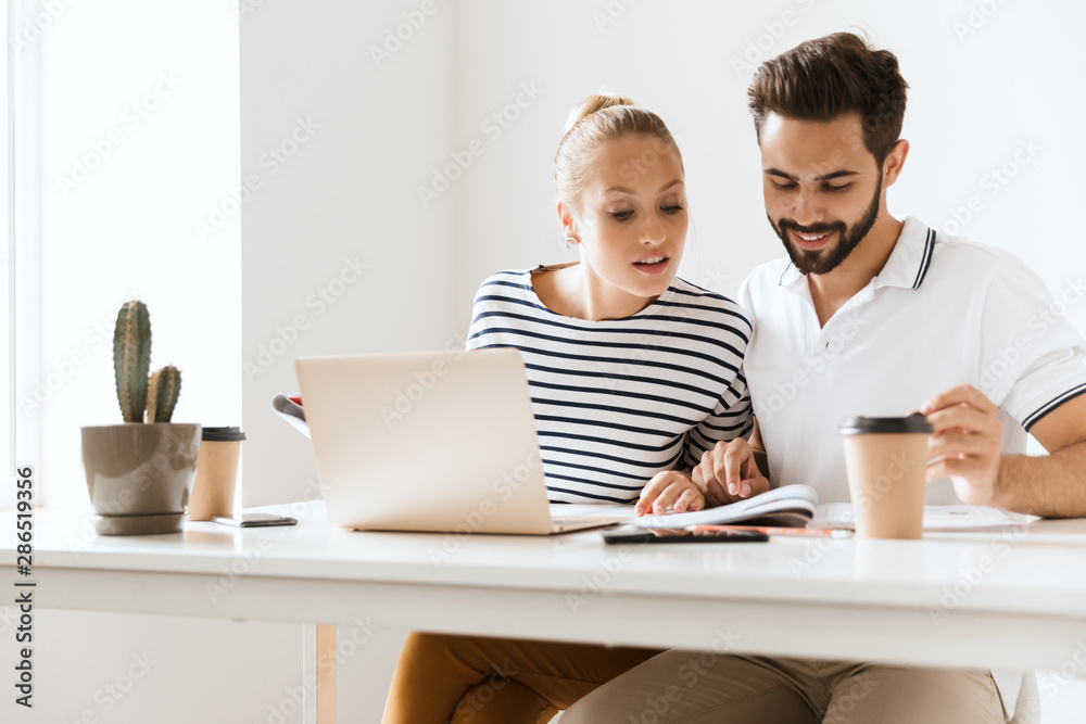 Optimistic happy pleased young loving couple friends man and woman sitting at the table indoors using laptop computer reading book studying. Optimistic happy pleased young loving couple friends man and woman sitting at the table indoors using laptop computer reading book studying.