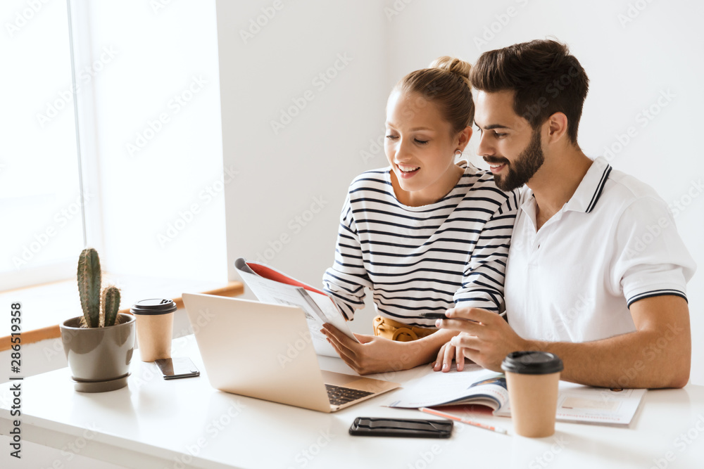 Optimistic happy pleased young loving couple friends man and woman sitting at the table indoors using laptop computer reading book studying. Optimistic happy pleased young loving couple friends man and woman sitting at the table indoors using laptop computer reading book studying.