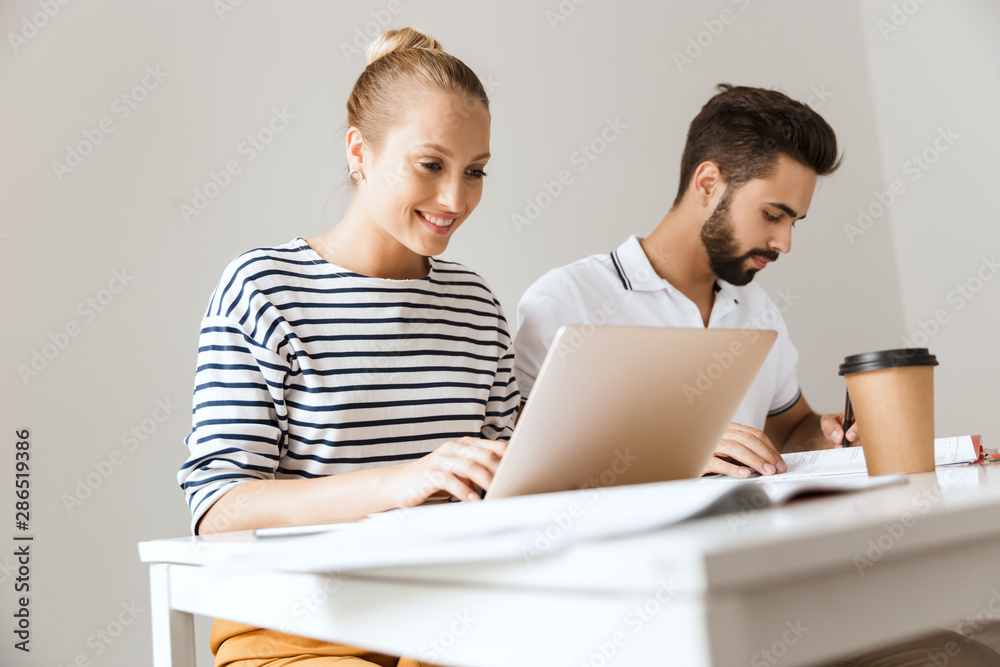 Cheerful loving couple friends man and woman sitting at the table indoors using laptop computer writing in notebook studying. Cheerful loving couple friends man and woman sitting at the table indoors using laptop computer writing in notebook studying.