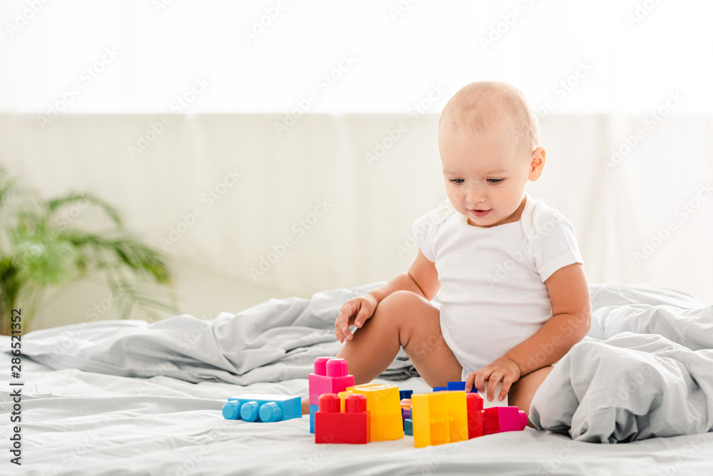 smiling baby in white clothes sitting on bed and looking at toys smiling baby in white clothes sitting on bed and looking at toys