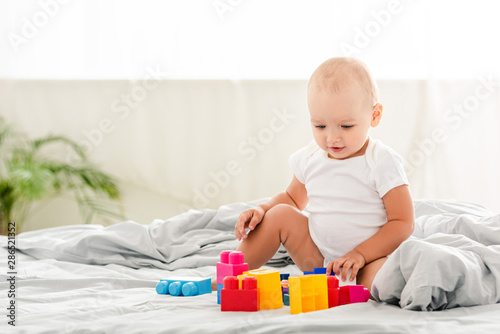 smiling baby in white clothes sitting on bed and looking at toys