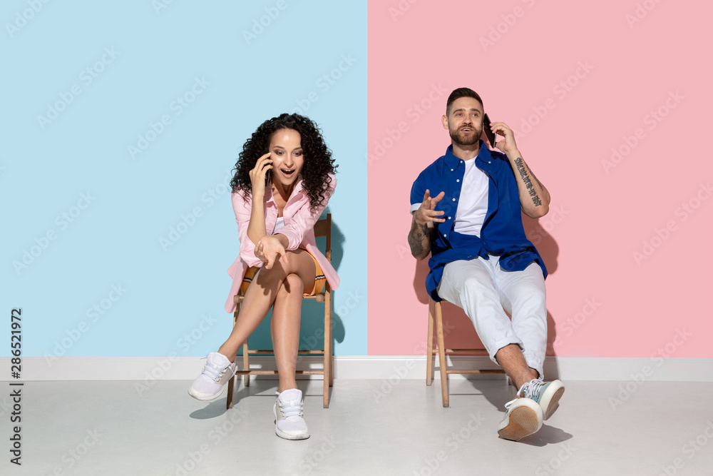 Naklejka premium Young emotional caucasian couple in bright casual clothes posing on pink and blue background. Concept of human emotions, facial expession, relations, ad. Woman and man talking on the phone, smartphone