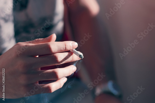 A  man s hand with slender fingers holds a Smoking cigarette  emitting blue smoke  illuminated by the light of the setting sun.