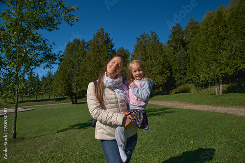 young blonde mother holding her toddler daughter in her arms as they are walking through the park on a sunny day © izikmd