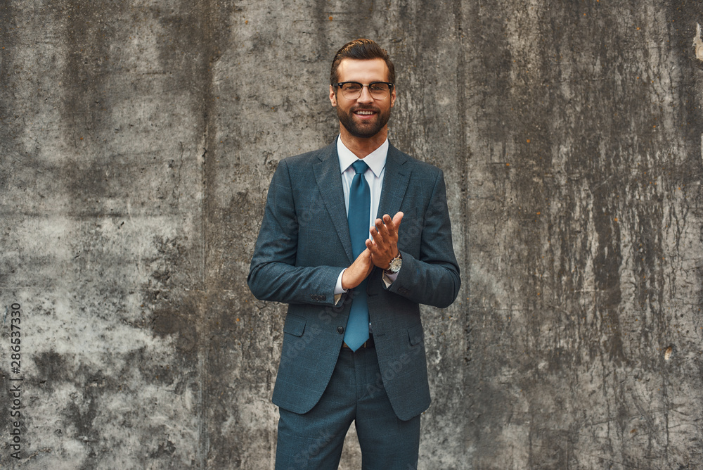 Success in everything. Confident and happy bearded businessman in full suit rubbing his palms and smiling while standing against grey stone wall outdoors Success in everything. Confident and happy bearded businessman in full suit rubbing his palms and smiling while standing against grey stone wall outdoors