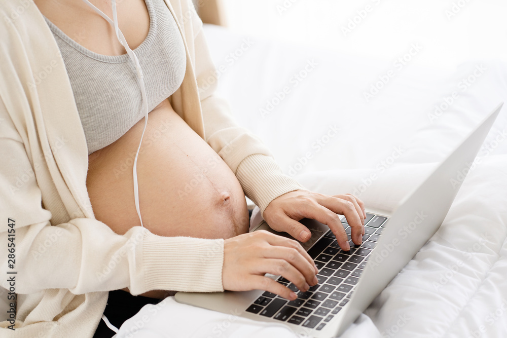 Naklejka premium Closeup at hands of Asian pregnant mother looking for music on Laptop will help to feel relax, happy and have a positive effect on unborn child. Online shop related to children accessories on social.