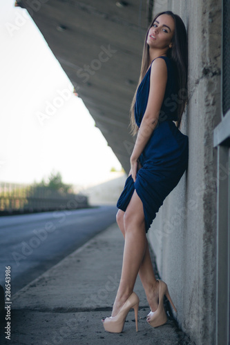 attractive woman in a long blue dress stands on her heels along a concrete wall. She has long hair, a belt and a pretty face.