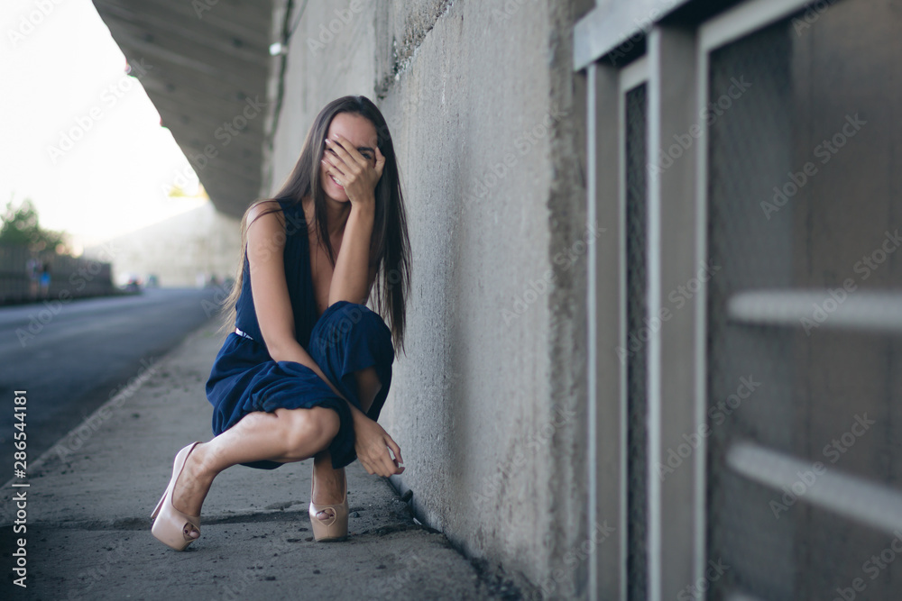 young elegant womana woman in a long dress squats on her heels against a long concrete wall stretching away into the distance. she smiles. cars on the background