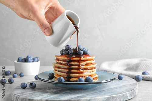 Woman pouring chocolate syrup onto fresh pancakes with blueberries at grey table, closeup