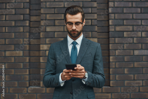Business message. Handsome businessman in formal wear and eyeglasses using smart phone while standing against brick wall
