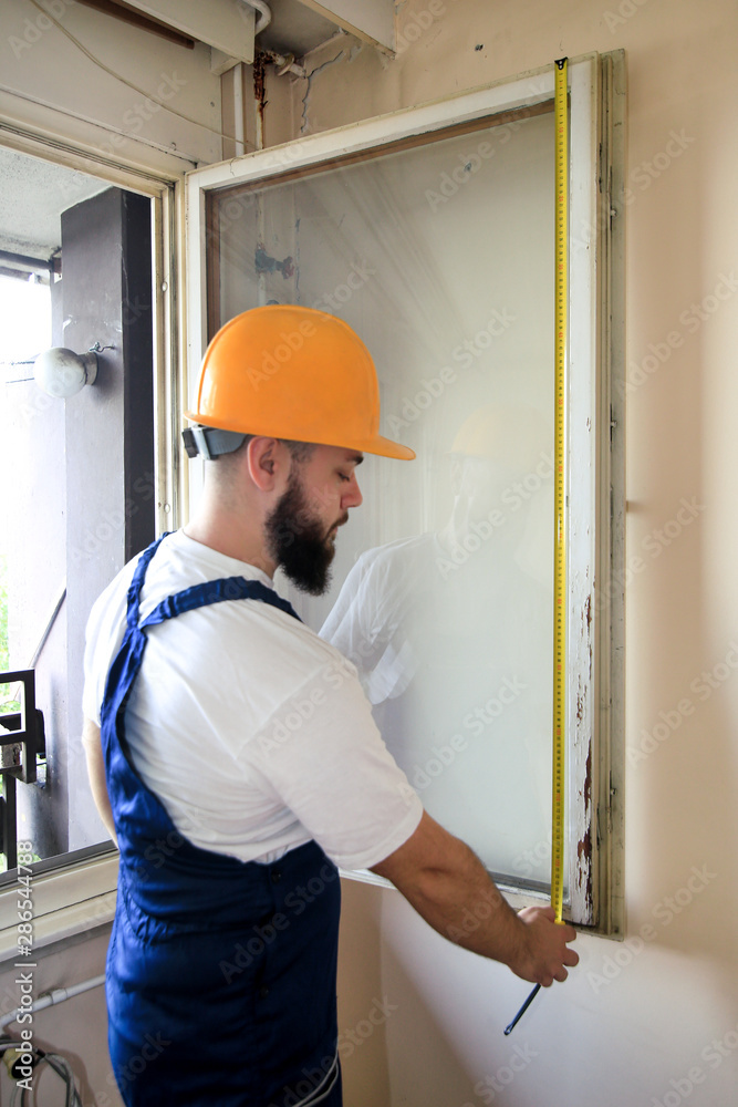 Construction worker and handyman is working on renovation of apartment. Builder is measuring window using measure tape at construction site. House renovation concept. Construction and service tools. Construction worker and handyman is working on renovation of apartment. Builder is measuring window using measure tape at construction site. House renovation concept. Construction and service tools.