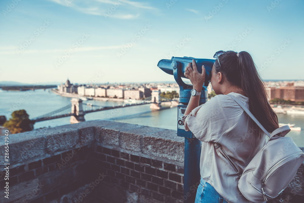 Obraz premium Pretty young brunette woman looking to historical city of Budapest by the big telescope, outlook, view to the city