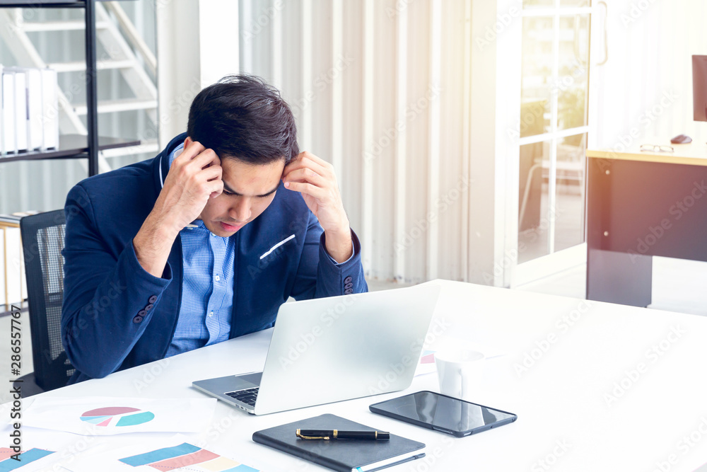 A young businessman sitting in a modern office. He has a feel stressed about the result of business profits not positive. On his table have a computer laptop tablet pen paper graph and a coffee cup.