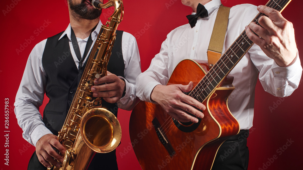 Fototapeta premium Group of two musicians, male jazz band, guitarist and saxophonist in classical costumes improvise on musical instruments in a studio on red background