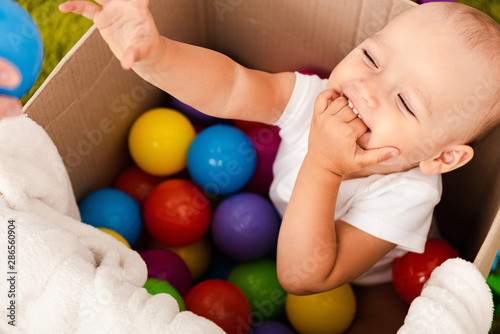 cute child sitting in cardboard box with colorful balls, laughing and raising his hand up