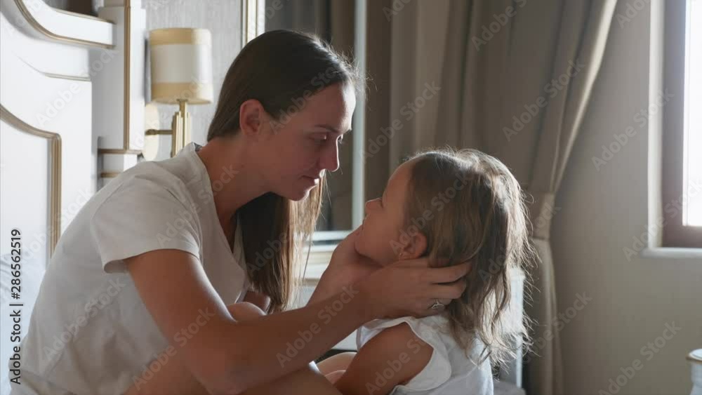 Young mother caresses her little daughter in bedroom at home. They sits on the bed together face to face, mom talks with child, embraces her and kissing. Young mother caresses her little daughter in bedroom at home. They sits on the bed together face to face, mom talks with child, embraces her and kissing.