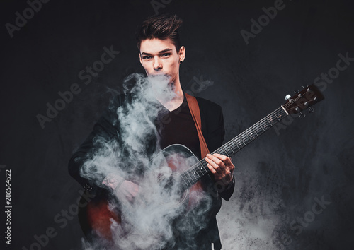 Cheeky young man is smoking, making nice vapour while holdind acoustic guitar at studio.