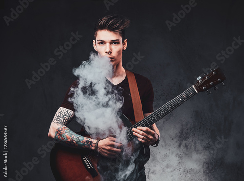 Young attractive man is playing acoustic guitar while smoking vape, making big haze.