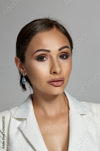 Close up portrait of a brunette model girl with professional evening make-up, wearing a white jacket with black bra, posing on gray background.