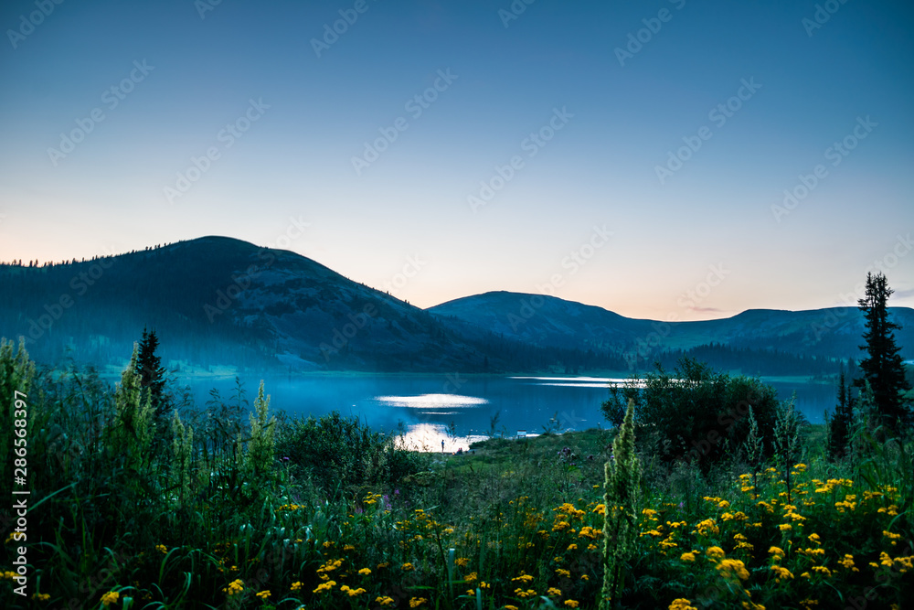 Beautiful mountain lake covered with smoke in front of hills and meadow with flowers in dusk, Ergaki, Krasnoyarsk region, Russia