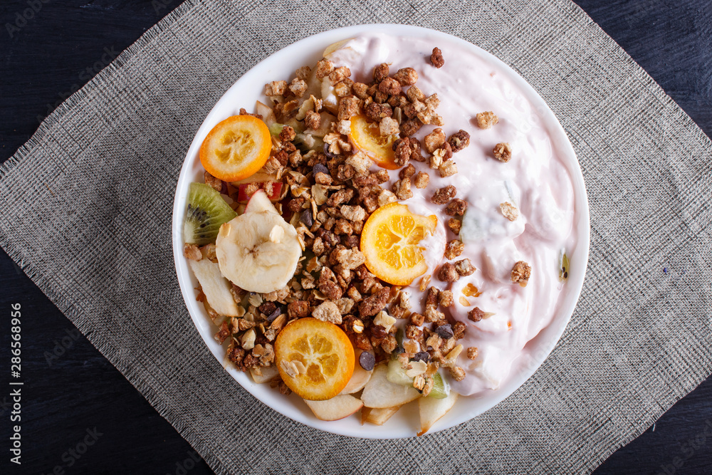 Vegetarian salad of bananas, apples, pears, kumquats, kiwi with granola and yogurt on black wooden background.