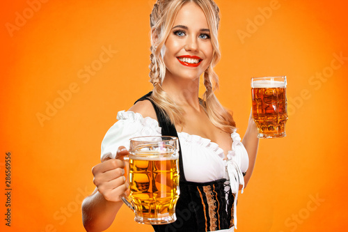 Young sexy oktoberfest girl waitress, wearing a traditional Bavarian or german dirndl, serving big beer mugs with drink isolated on yellow background.
