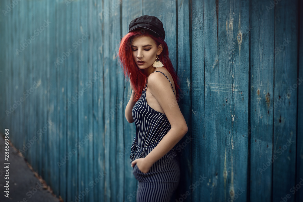 Fototapeta premium Young fashionable woman in clothes in a strip and red hair, posing on a blue background on the street.