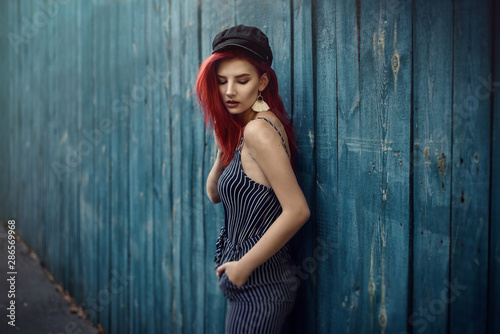 Young fashionable woman in clothes in a strip and red hair, posing on a blue background on the street. © Natalia