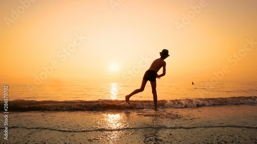 Young boy with hat skips stones by the sea beach at sunset, SLOW MOTION