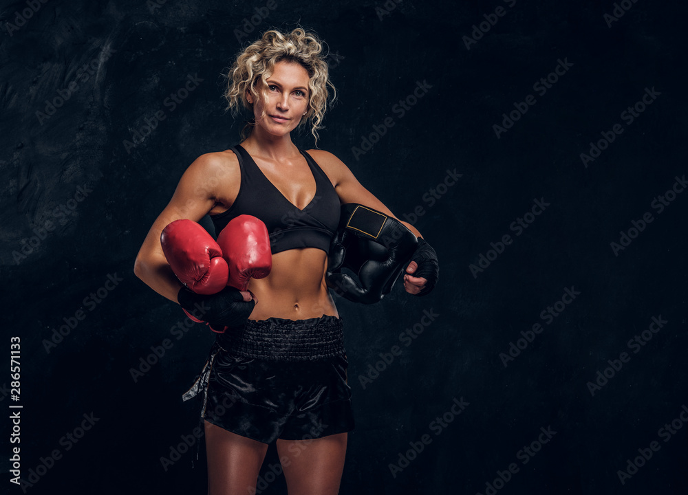 Experienced female boxer is posing for photographer at dark photo studio with equipment in her hands.