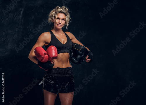 Experienced female boxer is posing for photographer at dark photo studio with equipment in her hands. © Fxquadro