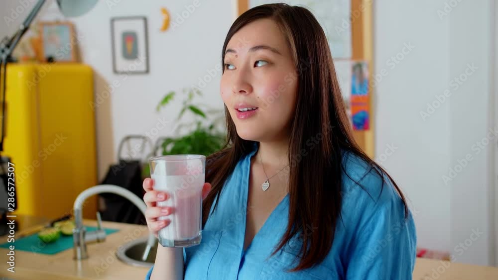 Alluring pensive Asian young woman drinking tasty fruit smoothie with coco milk at kitchen, cute smiling female in blue shirt with long brunette hair enjoying fresh yellow fridge on background Alluring pensive Asian young woman drinking tasty fruit smoothie with coco milk at kitchen, cute smiling female in blue shirt with long brunette hair enjoying fresh yellow fridge on background