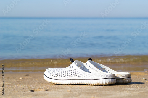 Holidays at sea background with copy space. Close-up of white sea slippers on the golden sand near the sea water.