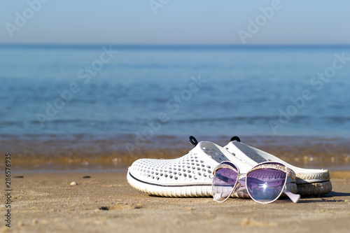 Recreation at sea background with copy space. Close-up of white sea slippers and sunlasses on the golden sand near the sea water.