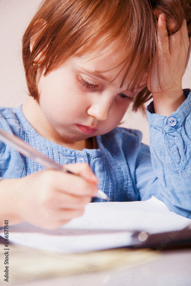 Tired and sad little child girl learning and writing writes in a workbook. Concept of difficult path to success and victory in the future. Tired and sad little child girl learning and writing writes in a workbook. Concept of difficult path to success and victory in the future.