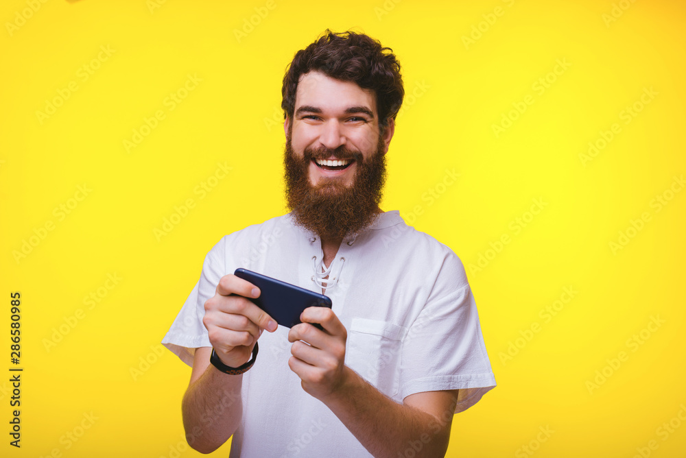 Cheerful bearded man playing or texting at his phone while looking at the camera smiling. Cheerful bearded man playing or texting at his phone while looking at the camera smiling.