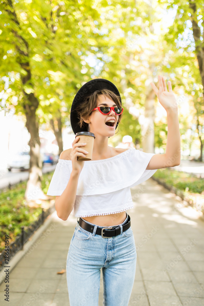 Gesture, lifestyle and people concept. Happy young woman or teenage girl drinking coffee from paper cup and wave hello on city street Gesture, lifestyle and people concept. Happy young woman or teenage girl drinking coffee from paper cup and wave hello on city street