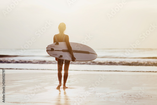 Surf girl with long hair go to surfing. Young surfer woman holding blank white short surfboard on a beach at sunset or sunrise.
